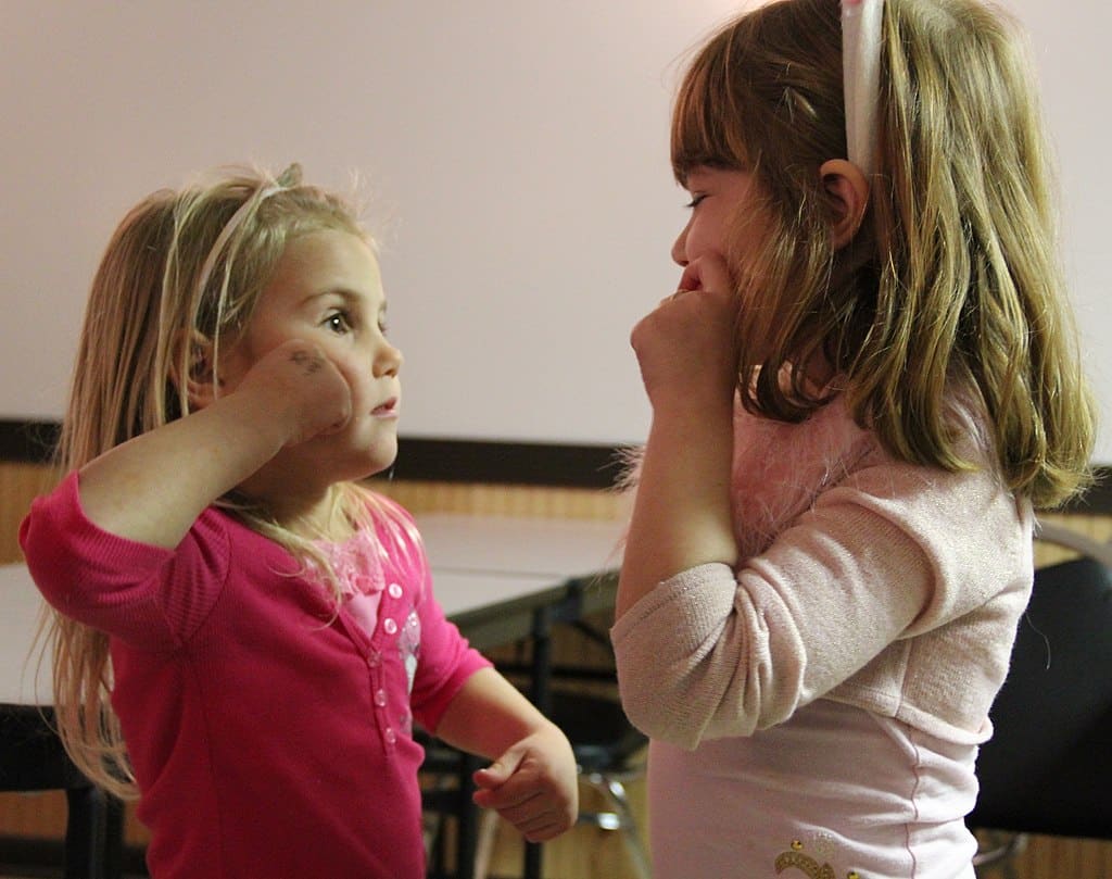 Kaksi 3–5-vuotiasta lasta viittovat toisilleen yhden käden viittomaa, jossa käsi tekee liikettä poskella kiertoliikkeellä.