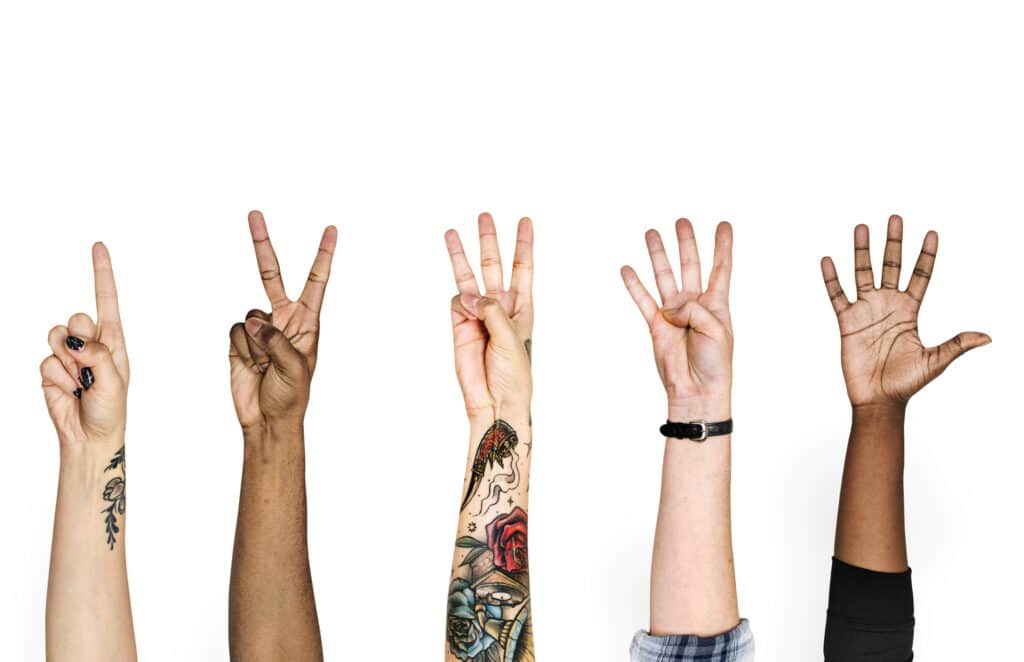 Neljän henkilön oikea käsi kurottuu ylöspäin ja elehtii numerot yhdestä neljään. Tausta on valkoinen. Kädet ovat erilaisia esimerkiksi ihonsävyltään ja tatuoinneiltaan.